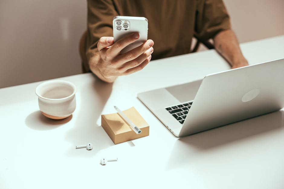 A person working remotely with a smartphone, laptop, and wireless earbuds on a desk.